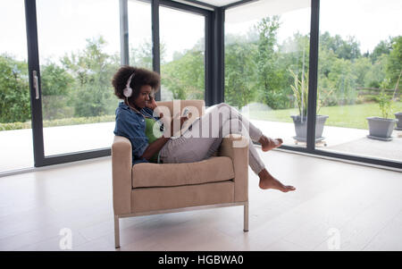 junge schöne afroamerikanische Frau hört gerne Musik mit Kopfhörern und Tablet in Ihrem Sessel in ihrem Luxushaus Stockfoto