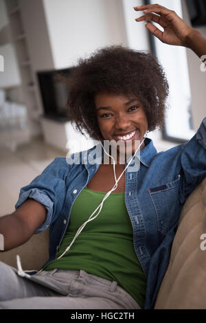 junge schöne afroamerikanische Frau hört gerne Musik mit Kopfhörern und Tablet in Ihrem Sessel in ihrem Luxushaus Stockfoto