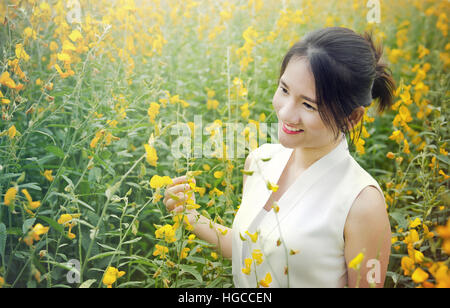 Porträtfoto des asiatischen Thai Mädchen mit Smiley-Gesicht und rote Lippe im Garten mit Morgenlicht und gelbe Sunhemp (Crotalaria Juncea) Blume ba Stockfoto