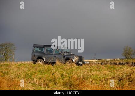 Ein langer Radstand Land Rover Defender fahren auf einer Landstraße B im Herbst mit einem dunklen Himmel hinter und grobe Wiese vor Stockfoto