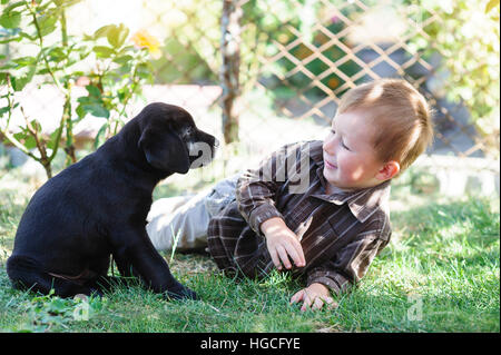 kleiner Junge mit einem schwarzen Labrador-Welpe liegt im Sommer Gras Stockfoto