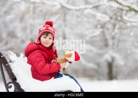 Entzückende kleine Kind, junge, spielen in einem verschneiten Park holding Teddybär, sitzen auf Bank, Winter Stockfoto