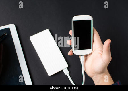 männliche Hände halten ein weißes Handy verbunden, eine Powerbank auf schwarz Stockfoto