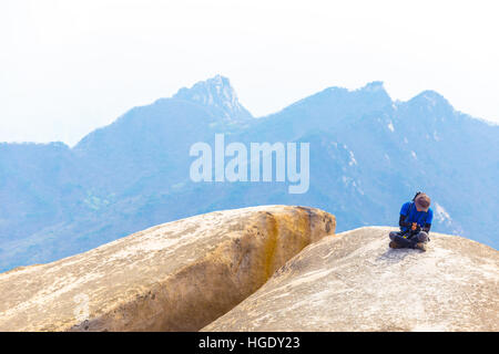 Koreanische Mann in bunten trekking Moden ruht auf Boulder auf Baegundae Gipfel nach dem Wandern Berg Bukhansan geschmückt Stockfoto