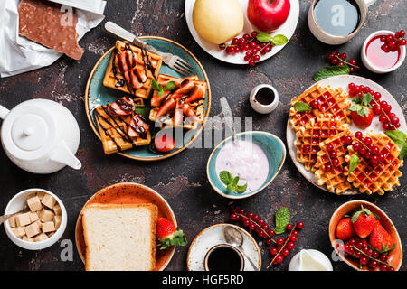 Frühstückstisch mit Waffeln, Joghurt, Kaffee und frisches Obst und Beeren auf schwarzem Hintergrund, Ansicht von oben Stockfoto