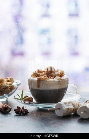 Glastasse Kaffee mit Schlagsahne, Karamell Popcorn, Zimt und Schokolade in der Nähe des Fensters Stockfoto