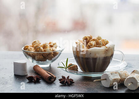 Glastasse Kaffee mit Schlagsahne, Karamell Popcorn, Zimt und Schokolade in der Nähe des Fensters Stockfoto