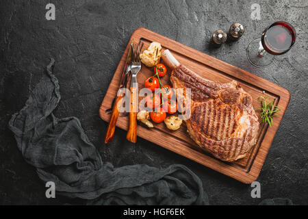 Saftig gegrillte Tomahawk Rindersteak mit Knochen mit Gewürzen, frischem Rosmarin und gegrilltem Gemüse auf einem schwarzen Hintergrund, Ansicht von oben Stockfoto