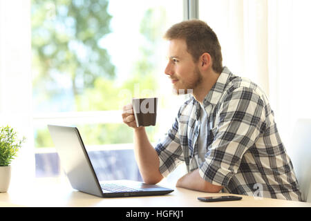 Nachdenklicher Mann mit einem Laptop, Blick durch ein Fenster hält eine Kaffeetasse zu Hause Stockfoto