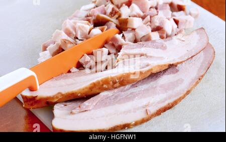 Nahaufnahme der Speckscheiben und Würfel und orange Messer auf Schneidebrett. Stockfoto