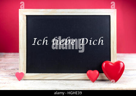 Hölzerne Tafel mit roten Herzen und geschriebenen Satz auf Deutsch "Ich Liebe Dich", das heißt 'Ich liebe dich', in rotem Hintergrund. Stockfoto