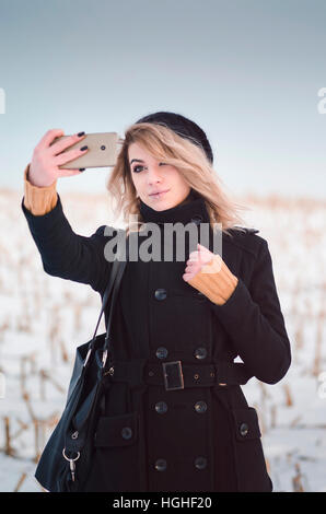 Junge alternative Blondine in schwarzer Kleidung nehmen Selfie im verschneiten Feld Stockfoto