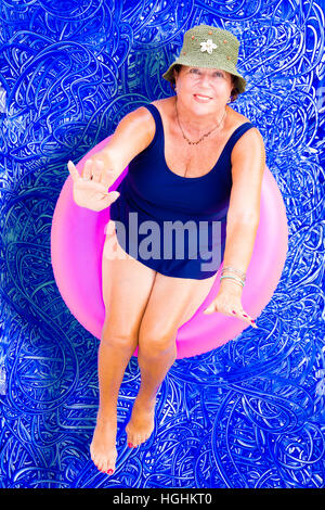 Ältere Dame in einem stilvollen Hut und Badeanzug schwimmend auf einem Rohr in das Schwimmbad entspannen Sie sich auf einen heißen Sommertag nachschlagen zu winken in die Kamera, Konz Stockfoto