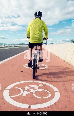 ein junger kaukasischen Mann Sport Kleidung gesehen von hinten mit seinem Fahrrad durch ein Radweg direkt am Meer Stockfoto
