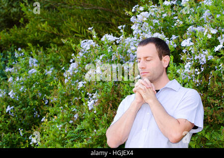 Mann, der betet allein außen mit Hände gefaltet vor einem Busch mit hellblauen Blüten. Stockfoto