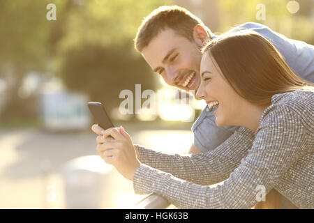 Seitenansicht eines Paares lachend beobachten Medieninhalte in ein smart Phone im Freien in einem Balkon bei Sonnenuntergang mit warmem Licht