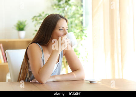 Porträt eines Mädchens, sitzend auf einem Stuhl, Kaffee zu trinken und auf der Suche nach außen durch ein Fenster zu Hause mit einem warmen natürlichen Licht Stockfoto