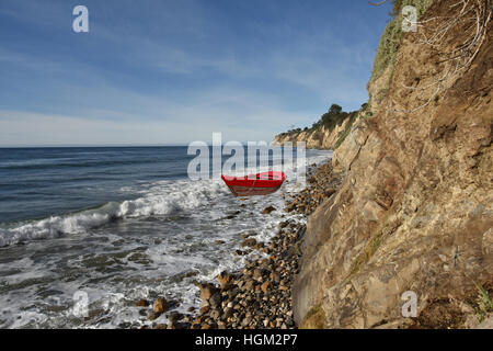 Rot Ruderboot in der Brandung an einem steinigen Strand unter einer Klippe. Stockfoto