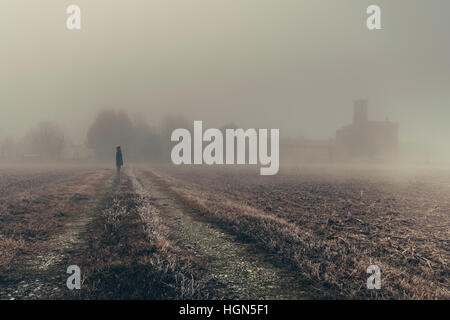 Frau in der Ferne Blick auf eine Kirche im dichten Nebel von der Region Lombardei in Italien während der Winter, gefrorenen Mais-Feld Stockfoto