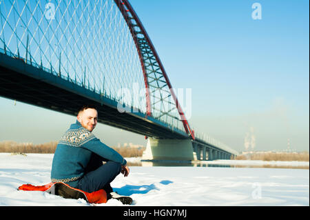Mann in warme Kleidung mit einem Bart ist auf der Straße im Winter in einem warmen, sonnigen Tag an der Flussbrücke im Hintergrund laufen, sitzt im Schnee Stockfoto