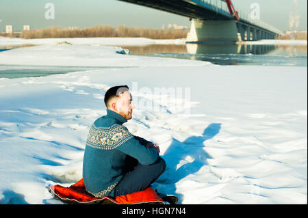 Mann in warme Kleidung mit einem Bart ist auf der Straße im Winter in einem warmen, sonnigen Tag an der Flussbrücke im Hintergrund laufen, sitzt im Schnee Stockfoto