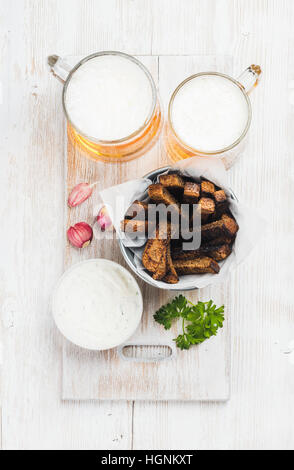 Bier-Snack-Set. Zwei Tassen Pilsener, Roggen Brot Croutons, Knoblauch-Käse-Sahne-Sauce und frischem Kraut über weiß bemalte alte hölzerne backgroundwith Stockfoto