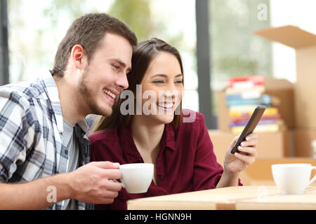 Liebevolle paar entspannen und mit einem Smartphone sitzen auf dem Boden im Innenbereich während der Bewegung Apartment mit Kartons im Hintergrund Stockfoto