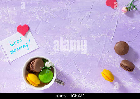 Frühstück, Muttertag, Geburtstag Hintergrund. Becher Kuchen Macaron, Blatt Notebook Inschrift guten Morgen auf lila Tischplatte. Kopieren Sie Raum. Stockfoto