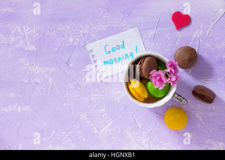 Frühstück, Muttertag, Geburtstag Hintergrund. Becher Kuchen Macaron, Blatt Notebook Inschrift guten Morgen auf lila Tischplatte. Kopieren Sie Raum. Stockfoto