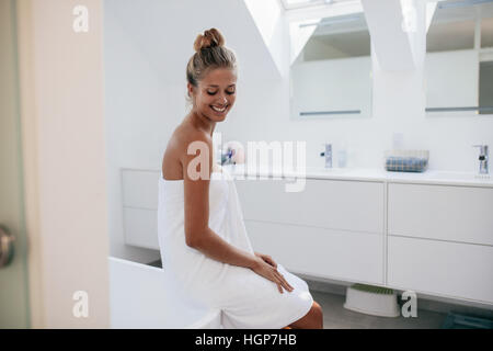 Lächelnde junge Frau sitzt auf Badewanne im Badezimmer nach Bad. Junge weibliche Modell in Handtuch gewickelt. Stockfoto