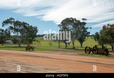 Alte rostige Landmaschinen neben einem roten Sandweg mit ländlichen treed Ackerland unter blauem Himmel mit Wolken in Western Australia. Stockfoto