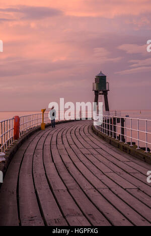 Mann allein zu Fuß auf Whitbys West Pier Ausdehnung unter einem dramatischen, lebhaftes, rosa, rote Himmel bei Sonnenuntergang - North Yorkshire, England. Stockfoto