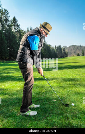 Senioren spielt Golf. Aktiven Ruhestand. Ein Mann ist Golfen, um in Form zu bleiben. Auf dem grünen Rasen mit Wald im Hintergrund Stockfoto