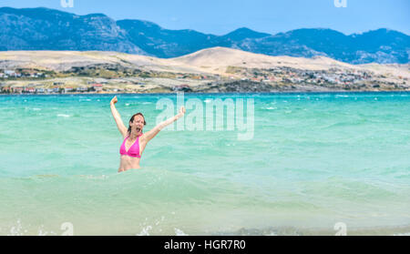 Frau springt aus dem Wasser, Lächeln, gesunde Lebensweise zu feiern. Arme gestreckt im blauen Meer mit Insel im Hintergrund. Stockfoto