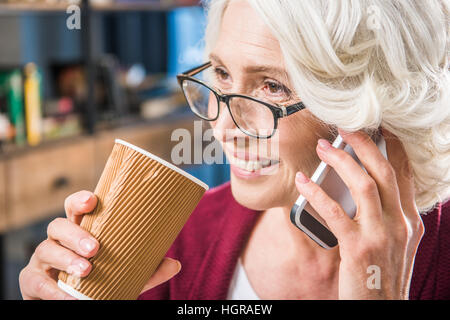 Attraktive senior Frau in Brillen am Smartphone sprechen und hält Pappbecher Stockfoto