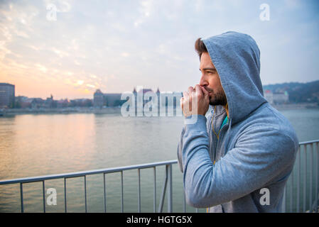 Ein hübscher junger Mann bläst seine Hände beim stehen in den Sonnenuntergang in Trainingskleidung Stockfoto