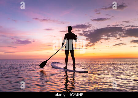 Person Stand-up-Paddle-boarding in der Abenddämmerung auf einem flachen warmen ruhigen Meer mit schönen Sonnenuntergang Farben Stockfoto