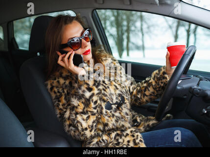 Schöne Frau in einem Pelzmantel mit roten Lippen Multitasking während der Fahrt, Kaffee trinken und reden am Telefon Stockfoto