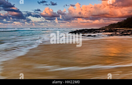 Seascape Sonnenaufgang über 75 Mile Beach auf Fraser Island in QUeensland, Australien. Schwarzen vulkanischen Granit Steinen Stand gegen Pazifik surft und Gezeiten an Stockfoto