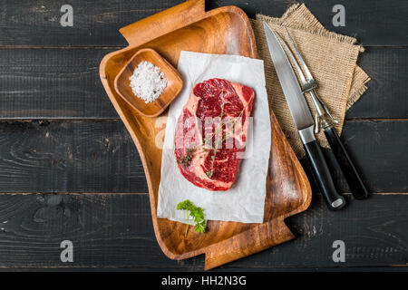 Rohes Frischfleisch Ribeye Steak, Gemüse, Salz und Thymian auf Schneidebrett mit Fleischgabel auf dunklem Holz, Ansicht von oben Stockfoto