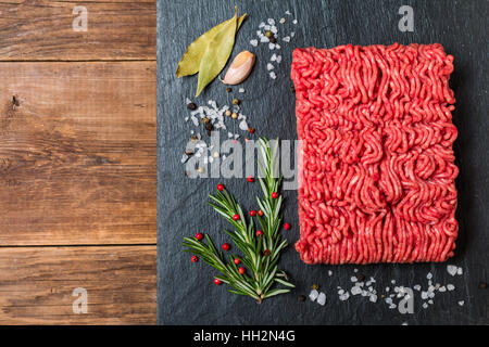 Hackfleisch / Faschiertes auf einer Schiefertafel Stein Tafel mit Gewürzen und frischen Rosmarin auf hölzernen Hintergrund, Ansicht von oben Stockfoto