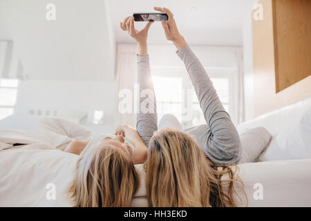 Mutter und Tochter am Bett liegen und unter Selbstbildnis mit Smartphone. Frau unter Selfie mit einem kleinen Mädchen im Schlafzimmer. Stockfoto