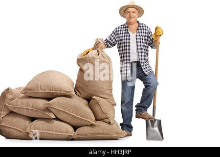 In voller Länge Portrait eines glücklichen älteren Bauern mit einer Schaufel stand neben einem Haufen von Sackleinen Säcke isolierten auf weißen Hintergrund Stockfoto