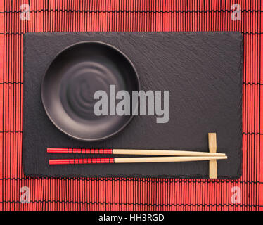 Ess-Stäbchen und schwarze Platte auf Schiefer Bahn auf roten Scroll Bambusmatte. Japanisches Essen Hintergrund.  Draufsicht mit Textfreiraum Stockfoto