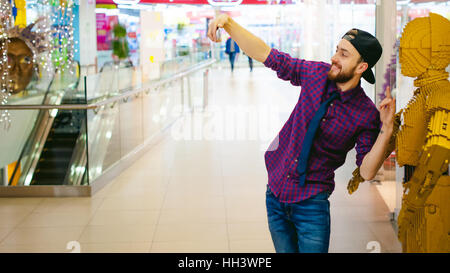 junger Mann mit einem Bart in ein kariertes Hemd und eine Krawatte und Cap, machen Selfie mit einem Filmhelden in der mall Stockfoto