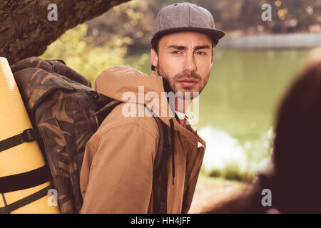 Junge bärtige Mann Reisenden mit Rucksack ansehen unter riesigen Baum in der Nähe von Fluss und Blick auf unscharfen Frau mit Pferdeschwanz im Vordergrund Stockfoto