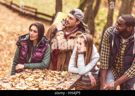 Gruppe von jungen Freunden Rucksacktouristen am Holztisch sitzend und wegschauen im herbstlichen Wald Stockfoto