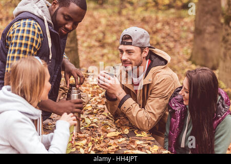 Gruppe von lächelnden Freunde sitzen am Tisch und gießt heißes Getränk im herbstlichen Wald Stockfoto