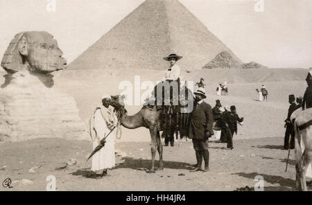 Touristischen Frau auf einem Kamel, begleitet von einem Mann und einem lokalen Führer in Gizeh, Ägypten. Im Hintergrund die große Sphinx und die Pyramiden von Gizeh zu sehen. Stockfoto