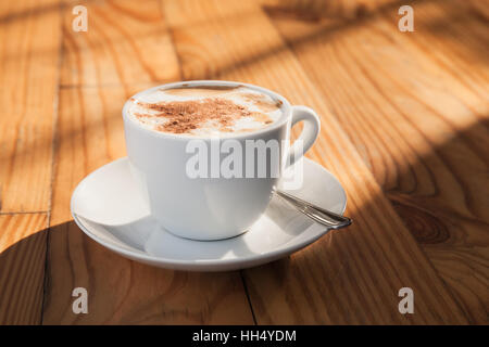 Cappuccino. Tasse Kaffee mit Milch Schaum und Zimt Pulver steht auf Holztisch im morgendlichen Sonnenlicht Stockfoto